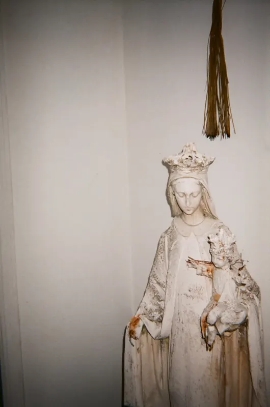 A weathered plaster statue of the crowned Virgin and Child against a white wall, a straw tassel hanging above