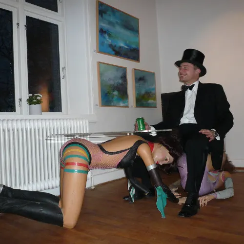 A man in a top hat and tailcoat seated by a glass table resting on two costumed women, in a gallery