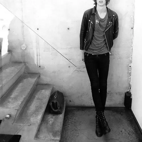 A thin young man in a leather jacket and black jeans beside a concrete staircase